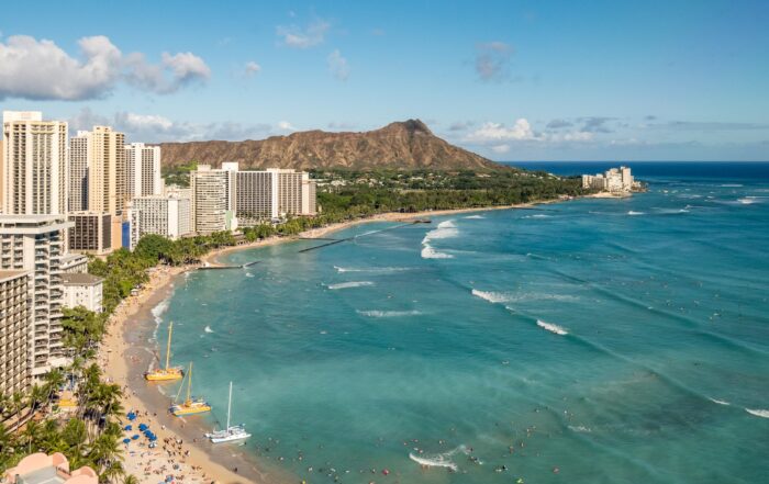 An aerial shot of Diamond Head. Waikiki beach stretches along the blue water. It’s a clear day. There are plenty of people on the beach and three sailboats are parked at the shore.