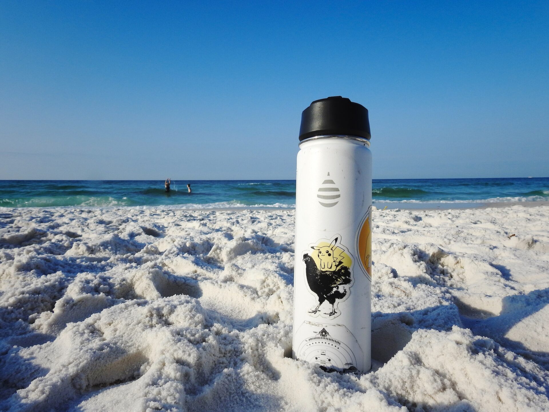A reusable water bottle with some stickers on it stands alone in the sand on a beach.The sand is pale and you can see the ocean just beyond it.