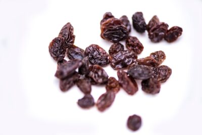 A handful of raisins are set on a white background. The raisins are dark purple and wrinkly. Some are stuck together. 