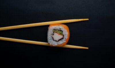 One piece of sushi is held between a pair of chopsticks against a black background. The sushi has fish on the outside, then a layer of rice, and more fish and cucumbers in the center. 