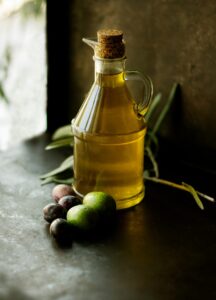 A full bottle of olive oil has been set on a dark green table. The bottle is surrounded by olives and has a stopper in its top. 