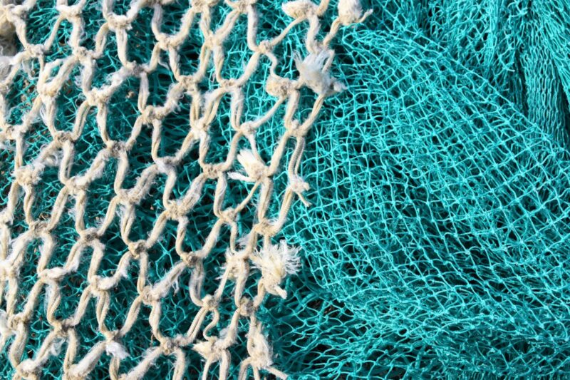A teal net with small holes takes up half the image. A beige, weathered, natural-looking net with larger holes takes up the other half. The beige net is laid on top of the teal one. Nets like these, if lost at sea, can result in ghost fishing as they’re made of materials that don’t biodegrade easily.