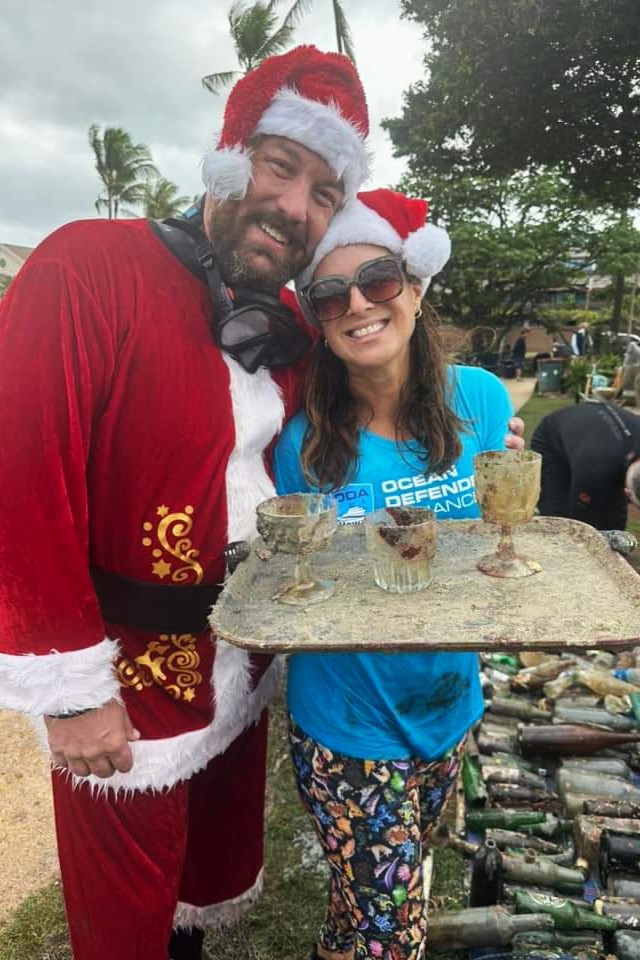 SCUBA Santa poses with Nudi Wear volunteer holding a plastic try and 3 glasses found while cleaning up the ocean