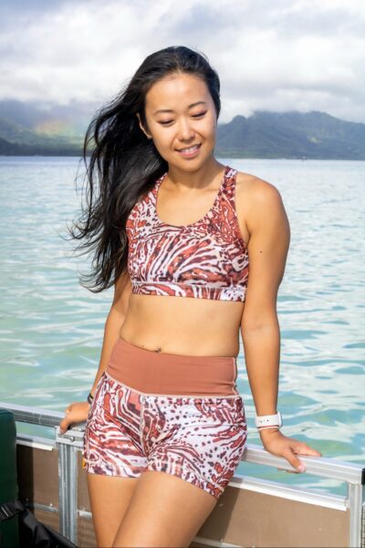 A model shows off Nudi Wear’s Lionfish Series gear. She’s wearing a swim top and shorts with the red and white lionfish patterns. Her hair is long and black, she has a belly ring, and has a white Apple watch on her left hand. She’s smiling softly and looking down. Behind her is the shallow blue water of Kaneohe Bay. In the distance you can see verdant mountains. 